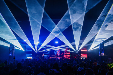 A large crowd at an covered concert with bright blue and white laser lights projecting over the audience, and colourful visuals displayed on screens behind the stage.