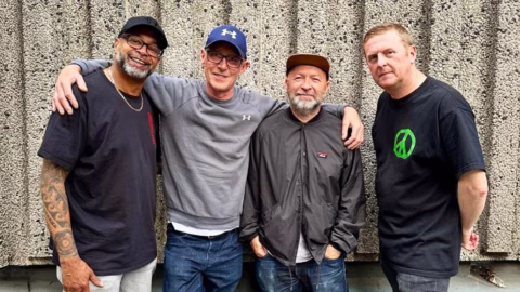 Four men stand closely together in front of a textured concrete wall, smiling and posing for the camera with their arms around each other. They are dressed casually in t-shirts, jackets, and caps.