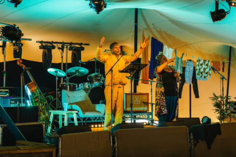 Two performers stand on a brightly lit stage. One raises both arms while speaking into a microphone, and the other gestures beside them.