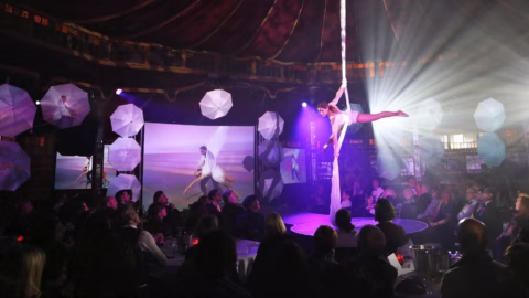 Aerial performer hangs from fabric high above a circular stage in a spiegeltent, watched by an audience seated at tables; spotlights and projections decorate the background.