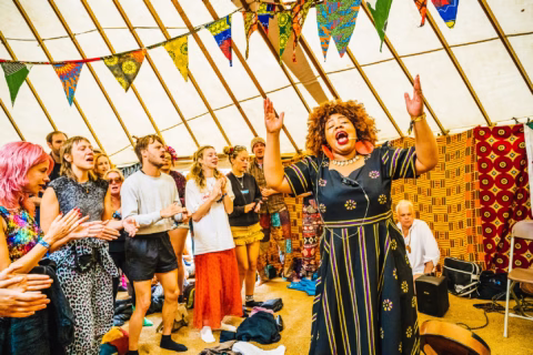 A joyful woman sings passionately with her arms raised while a diverse group of people stand around her, clapping and singing inside a colourful tent decorated with patterned flags.