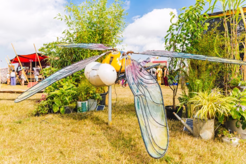 A large, colourful dragonfly sculpture with transparent wings is displayed outdoors among green plants at a festival, with people and tents visible in the background under a blue sky with clouds.