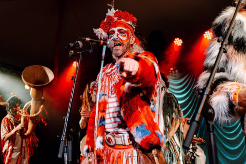 A performer in colourful, furry costume and face paint points towards the camera whilst singing on stage, surrounded by other similarly dressed musicians under dramatic lighting.