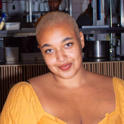 A person with a shaven head, wearing a yellow top, smiles at the camera inside a kitchen or restaurant setting with shelves and utensils in the background.