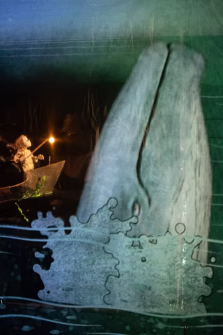 A puppet of a person in a small boat faces a large, illustrated whale emerging from the water, with splashes and waves around its head, under dim, blue-green lighting.