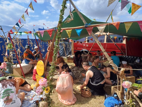 A group of people sit and work on crafts under a decorated marquee with colourful bunting at an outdoor festival. Flowers and plants adorn the area, and a red van is parked nearby under a partly cloudy sky.