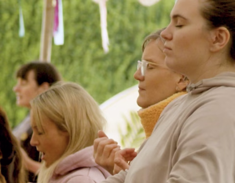 A group of women seated outdoors with their eyes closed, appearing calm and meditative. They are wearing casual clothing and facing the same direction, with greenery in the background.