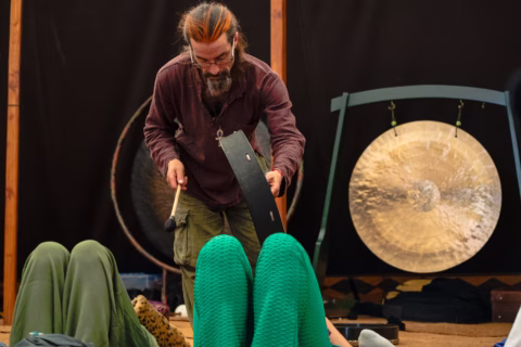 A man with long hair and a beard uses a drum over two people lying down, with a large gong in the background. The setting appears to be a relaxation or sound therapy session.