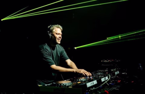 A DJ wearing headphones smiles whilst mixing music at a DJ booth, surrounded by equipment. Green laser lights shine through a dark background, creating a lively atmosphere.
