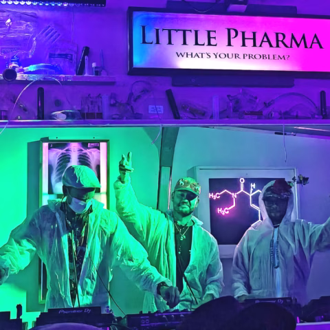 Three DJs wearing white lab coats and sunglasses perform under colourful neon lights in front of a bar sign that reads LITTLE PHARMA: WHAT'S YOUR PROBLEM? with scientific decor and a glowing molecular diagram behind them.