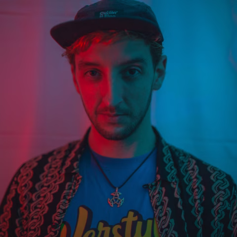A young person with short blond hair wears a dark cap, patterned shirt, and blue T-shirt, standing under red and blue lighting, looking directly at the camera with a neutral expression.