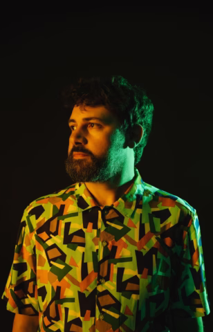 A man with a beard and moustache stands against a dark background, wearing a brightly coloured geometric-patterned shirt, illuminated by dramatic green and yellow lighting.