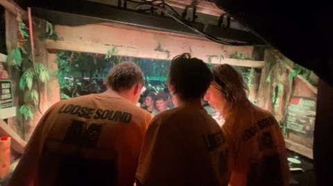 Three people wearing yellow LOOSE SOUND shirts stand side by side at a DJ booth, facing a crowd at a lively indoor event with leafy decorations and glowing lights.