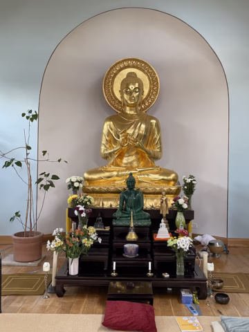 A golden Buddha statue sits in a shrine with a green Buddha figure below it, surrounded by flowers, offerings, candles, and spiritual items, set against a pale arched alcove in a serene room.