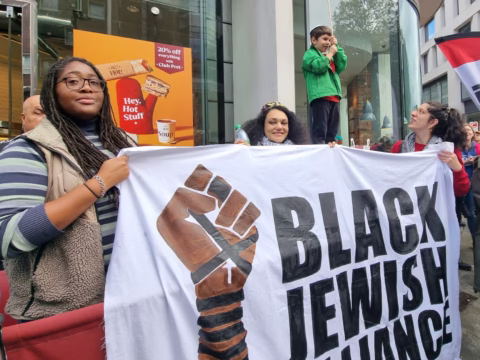 A diverse group holds a banner with a raised fist and the words Black Jewish Alliance at a protest. Behind them, a child in a green jacket speaks into a microphone while adults look on. A coffee shop is visible in the background.