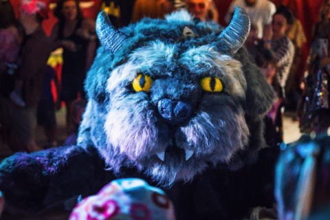 A person in a large, furry monster costume with yellow eyes, horns, and fangs moves through a crowd of people at a festive indoor event.