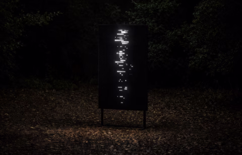 A black rectangular sign with small holes illuminated from behind, creating an abstract vertical pattern of light, stands in a dark outdoor area surrounded by trees and fallen leaves.