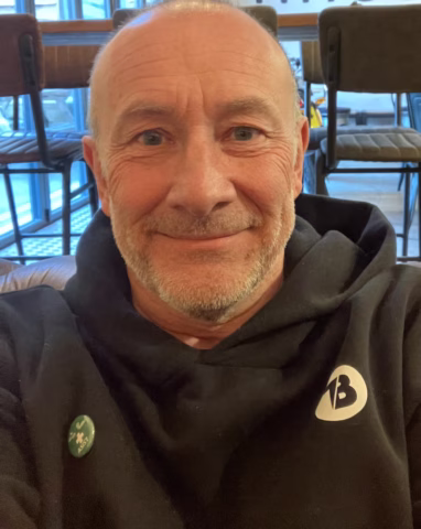 A smiling, middle-aged person with a shaved head and light stubble, wearing a black hoodie with a 13 logo and a small green badge, is sitting indoors in front of empty chairs and large windows.
