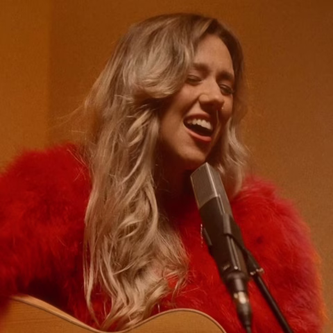 A woman with long, wavy blonde hair wearing a bright red, fluffy jumper sings passionately into a microphone while playing an acoustic guitar.
