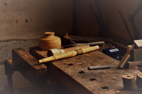 A wooden workbench with woodworking tools, a carved wooden spindle, a small wooden bowl, and other items, all bathed in soft, natural light in a rustic workshop setting.