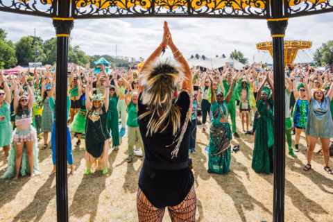 A woman leads a large outdoor crowd, all raising their arms in unison. Many people wear green costumes, and the scene appears festive, with tents and trees in the background on a sunny day.