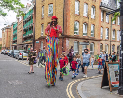 A performer on tall stilts dressed in colourful clothes and a red hat walks down a city street as a group of children and adults, some holding hula hoops, follow and watch. Brick buildings and parked cars line the road.