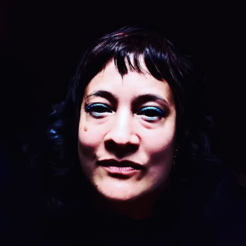 A person with short dark hair and blue eye make-up looks directly at the camera, illuminated by a strong light against a dark background.