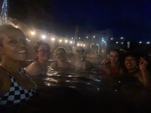 Six people are relaxing in an outdoor hot tub at night, surrounded by fairy lights. One person is holding a drink, and several are smiling at the camera. The atmosphere is warm and festive.