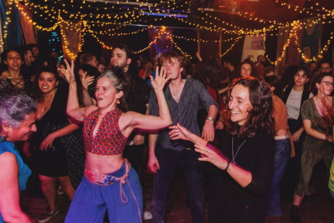 A lively crowd of people dance energetically under warm, yellow fairy lights in a festive indoor setting. Most are smiling and enjoying the music, creating a joyful and vibrant atmosphere.