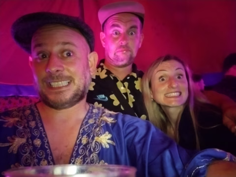 Three people pose for a selfie with wide, exaggerated smiles under pink-purple lighting. The man in front wears a blue, gold-embroidered shirt and a cap; the man and woman behind him appear happy and playful.