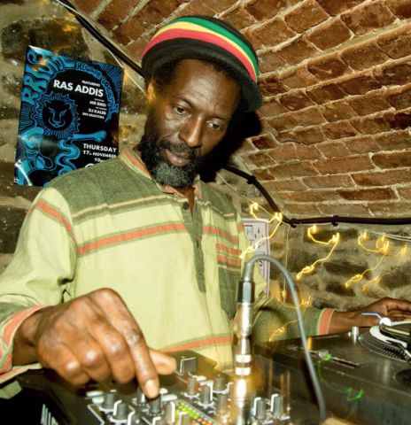 A man wearing a colourful striped hat and a beige shirt adjusts audio controls on DJ equipment under a brick arched ceiling. A poster on the wall behind him reads “Ras Addis.” Warm fairy lights decorate the stone wall.
