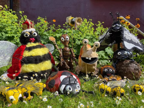 A group of colourful, handmade insect and bug sculptures made from various materials are displayed on grass with flowers and greenery in the background. The figures include a bee, ladybird, snail, ants, and other whimsical bugs.