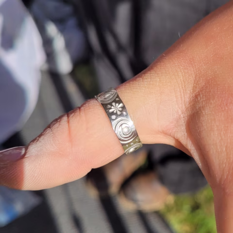 A close-up of a hand wearing a silver ring with engraved patterns, including swirls and a flower, on the thumb. The background is blurred with hints of grass and clothing.