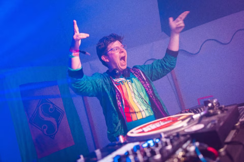A DJ wearing glasses, a colourful shirt, and a shiny jacket enthusiastically gestures with both hands while performing at a DJ booth, surrounded by vibrant lighting and equipment.
