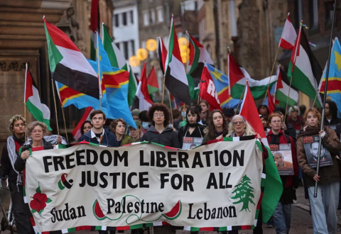 A group of people march holding flags and a large banner that reads Freedom Liberation & Justice For All: Sudan Palestine Lebanon during a protest. The crowd appears serious and focused.