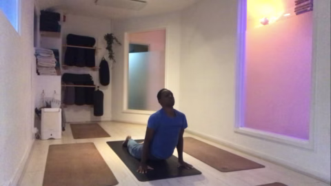 A person practises yoga alone in a bright, peaceful studio, performing the upward-facing dog pose on a mat. The room has stacked towels, yoga props, and warm, ambient lighting.