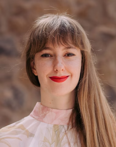 A woman with long light brown hair and fringe is smiling softly. She is wearing a light, floral-patterned top with a high collar and bright red lipstick. The background is blurred and earthy-toned.