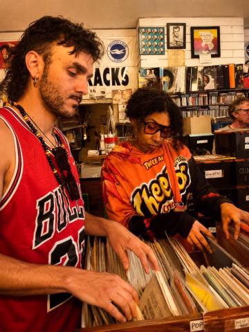 Two people browse through vinyl records in a music shop. One wears a red Chicago Bulls jersey, and the other wears an orange Cheetos hoodie. Shelves with CDs and music-themed decor fill the background.