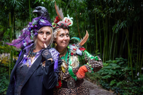 Two women in colourful, eccentric costumes and hats with feathers and flowers pose playfully in a lush garden with bamboo and greenery in the background. One woman holds a finger to her lips.