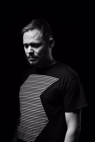 A man with short hair and a beard stands against a black background, wearing a black T-shirt with white horizontal stripes forming a geometric pattern. He looks downwards, and dramatic lighting highlights one side of his face.