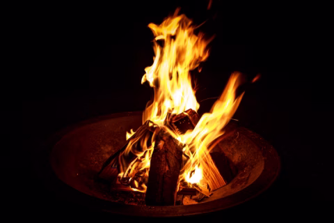 A campfire burns brightly in a firepit, with tall orange and yellow flames rising above stacked wooden logs against a dark background.