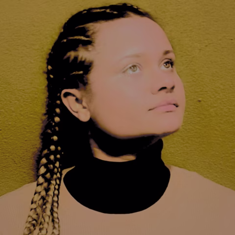 A person with plaited hair looks upwards, wearing a black polo neck and beige top, standing in front of a textured yellow-green wall.
