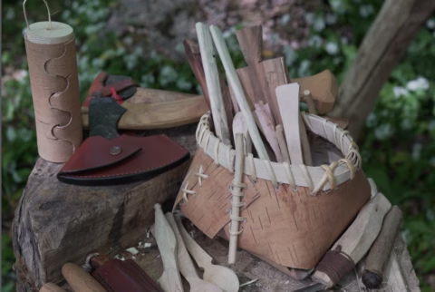 A woven basket filled with wooden utensils sits on a tree stump alongside axes, knives with leather sheaths, a birch bark container, and various carved tools. Green foliage is visible in the blurred background.