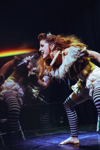 A barefoot performer in a theatrical, ruffled costume with striped leggings sings passionately into a microphone, leaning forward with expressive energy, reflected in a mirror behind her under dramatic stage lighting.