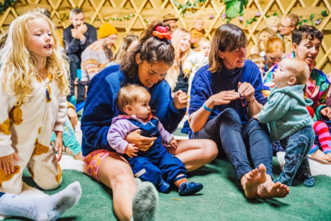 A group of adults and young children sit and play together on a green carpet in a cosy, brightly lit room. The atmosphere is joyful and relaxed, with people smiling and interacting closely.