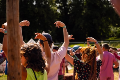 A group of people outdoors with their arms raised and hands bent at the wrists, appearing to dance or participate in an activity together; sunlight shines on them and trees are visible in the background.