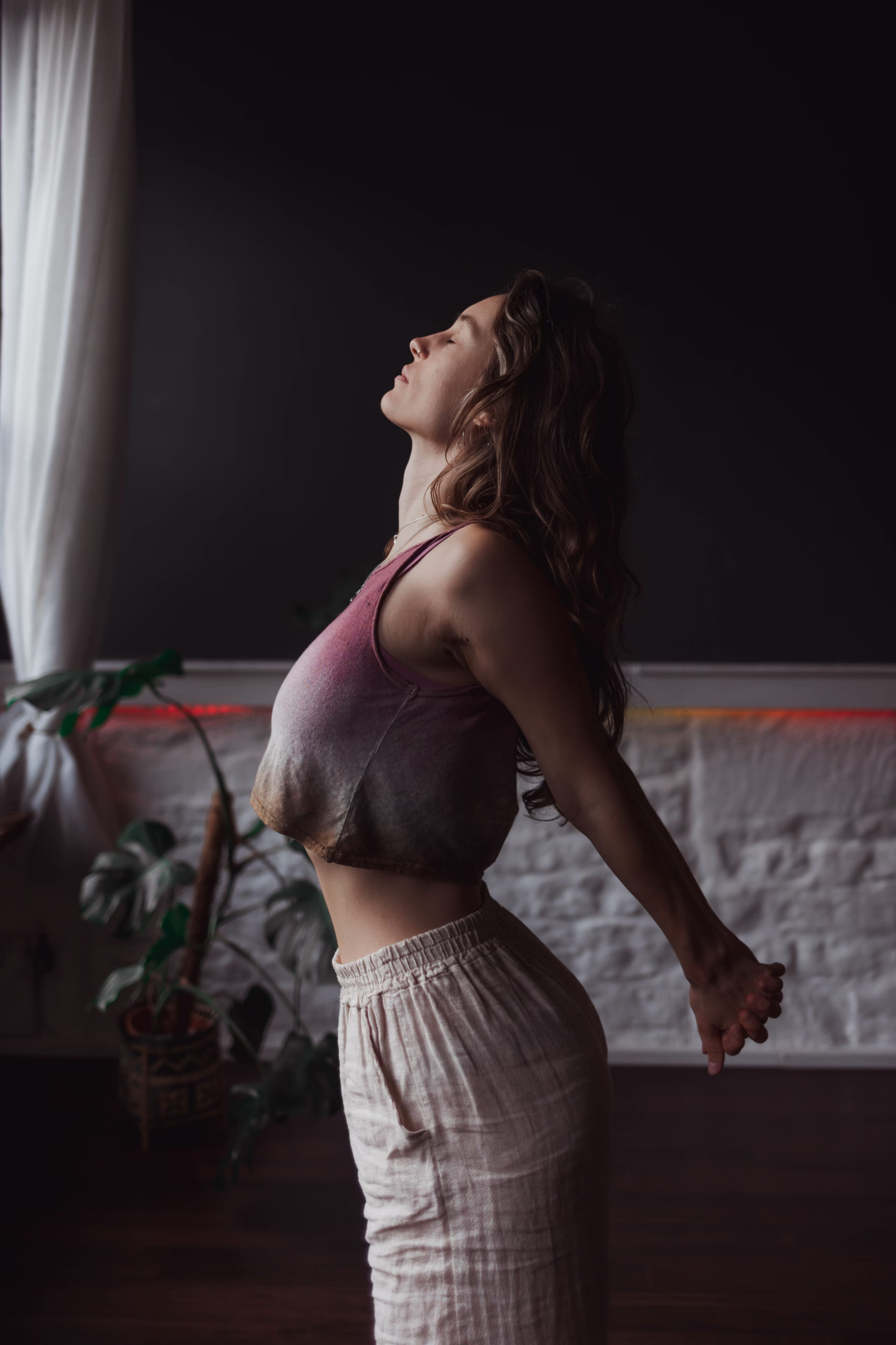 A woman in a vest and loose trousers stands indoors, stretching with her arms behind her back and head tilted upwards, eyes closed, near a window with light streaming in.