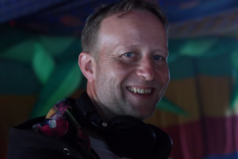 A smiling man with short hair wears headphones around his neck, standing indoors with colourful, blurred backgrounds.