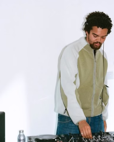A man with curly hair wearing headphones and a light-coloured jacket operates a DJ controller against a plain white wall. A water bottle is visible on the table beside him.