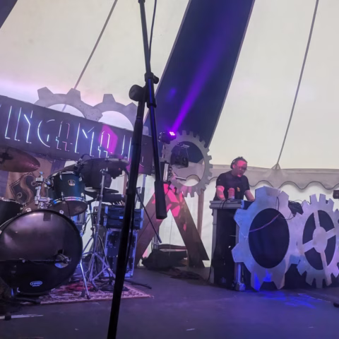 A musician wearing headphones performs on a keyboard behind large decorative gears on a stage, with a drum kit to the left and an Incandescena sign illuminated in the background under a marquee.